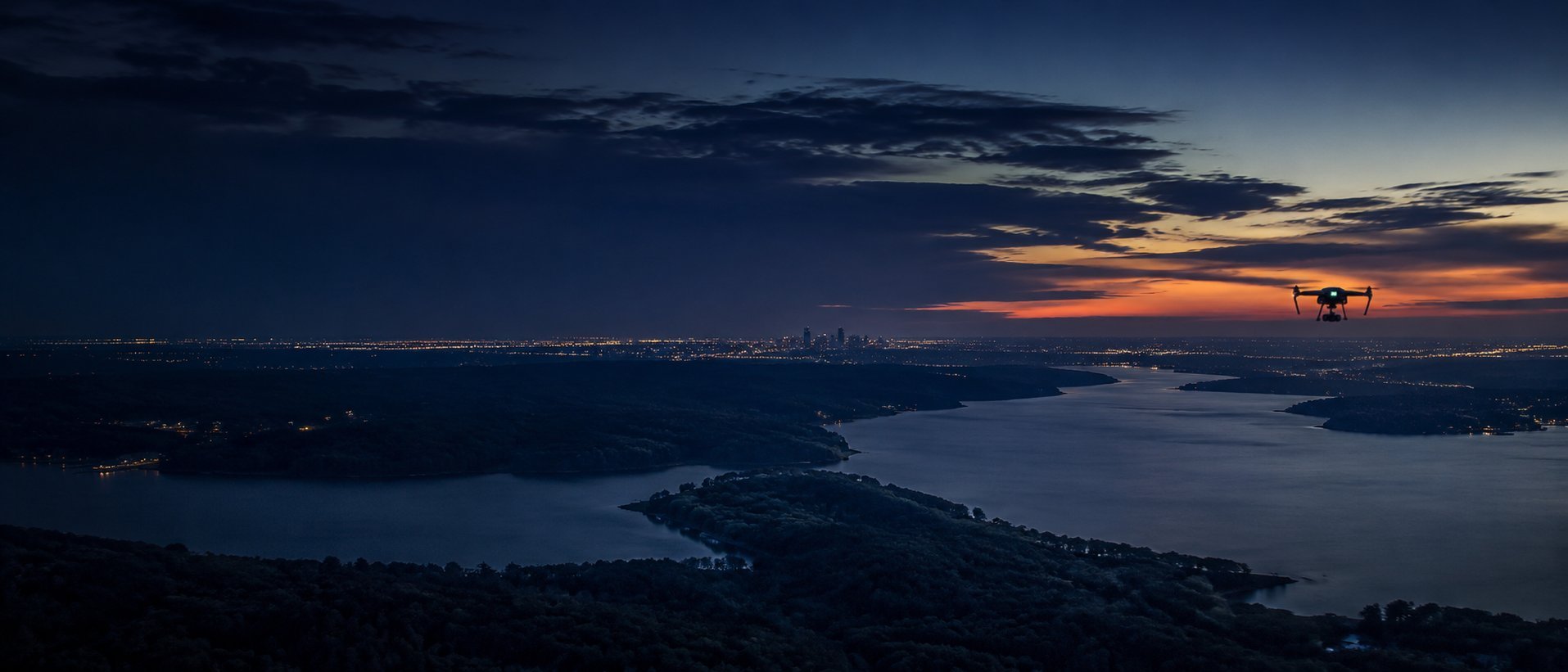 Aerial view of a lake surrounded by forested islands at twilight with a small aircraft silhouetted against an orange sunset sky