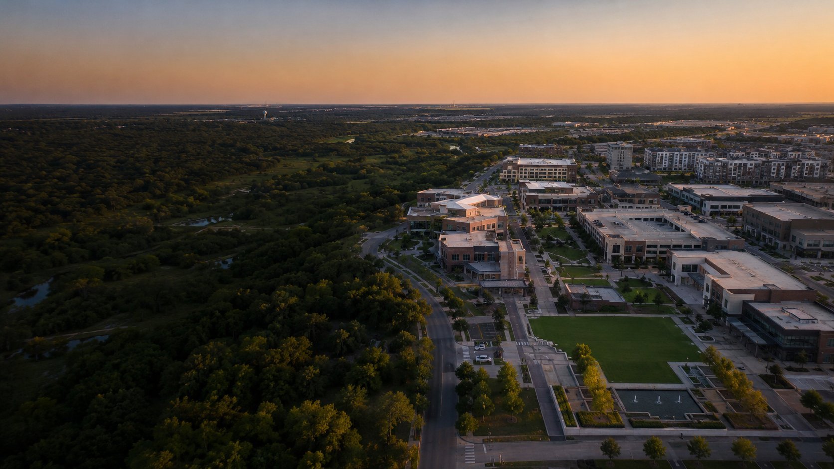 Aerial sunset view of suburban development with green forest, buildings, and roads transitioning to residential area
